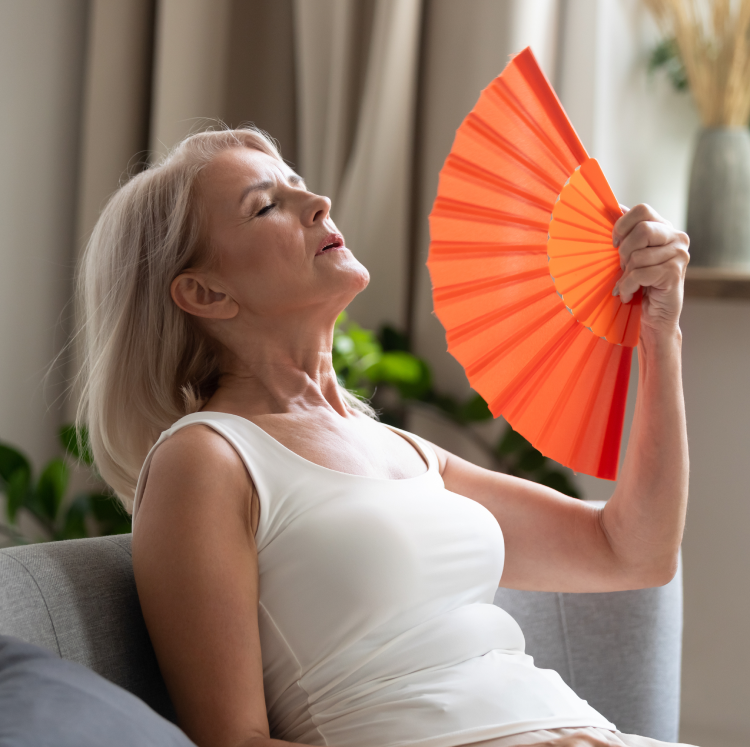 alt="Femme mûre se rafraîchissant avec un éventail orange, assise à l’intérieur et ressentant une bouffée de chaleur"