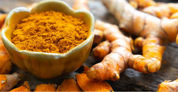 alt="Una ciotola piena di curcuma in polvere arancione vivace circondata da radici fresche di curcuma"