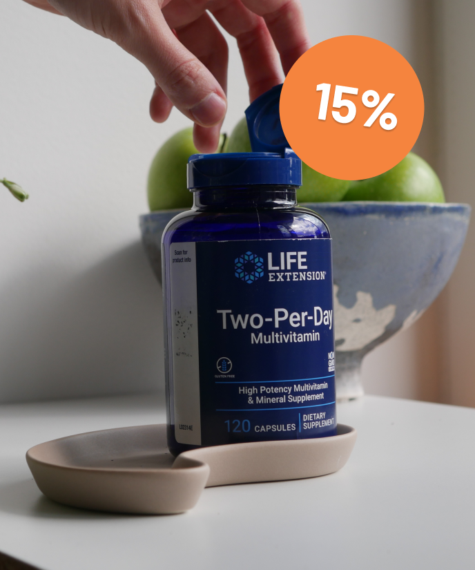 alt="Mano che apre un flacone di multivitaminico Two-Per-Day di Life Extension su una superficie bianca, con mele verdi sullo sfondo. Badge sconto del 15%."