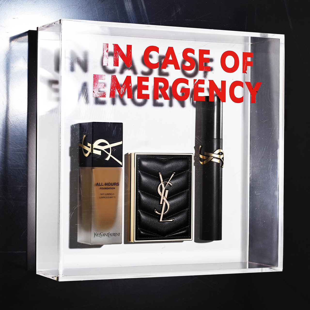 Prodotti di bellezza YSL, tra cui il fondotinta All Hours, una cipria compatta trapuntata e un mascara, esposti in una scatola trasparente con la scritta 'IN CASE OF EMERGENCY'.