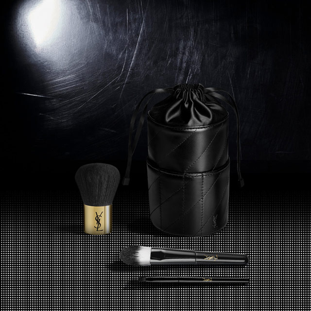 Set di pennelli da trucco YSL, inclusi un grande pennello per cipria e due pennelli più piccoli, accompagnati da un astuccio cilindrico nero trapuntato con il logo YSL.