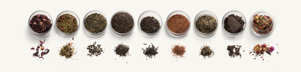 alt="Fila di piccole ciotole di vetro piene di vari tè sfusi, ciascuna con un piccolo mucchio di foglie di tè davanti, tra cui ibisco, tè verde, tè nero, rooibos e miscele floreali."