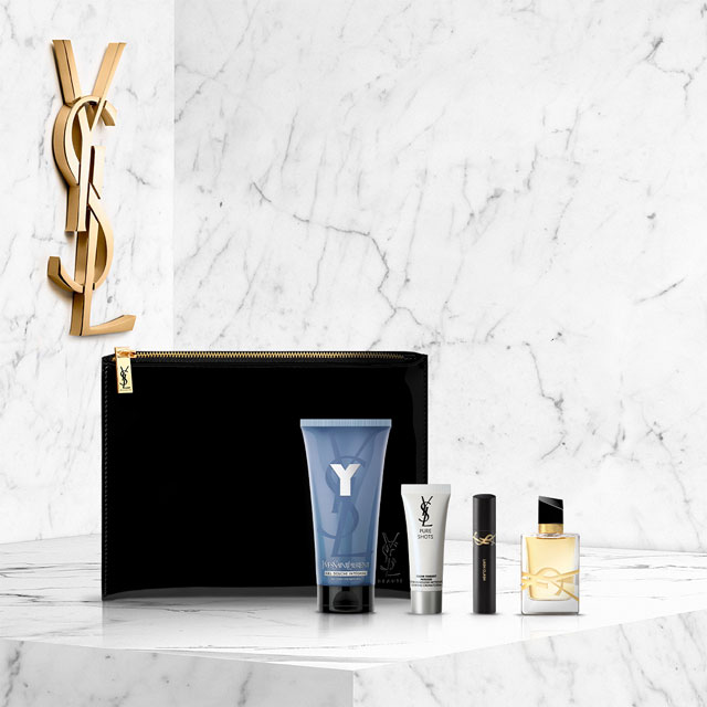 Un assortimento di prodotti di bellezza e profumi YSL da uomo, tra cui una borsa da toilette nera, un tubo di cura detergente, una piccola bottiglia "Pure Shots", un mascara o correttore e una bottiglia di profumo YSL Y. Il tutto è disposto su una superficie di marmo bianco con un grande logo YSL dorato in alto a sinistra.