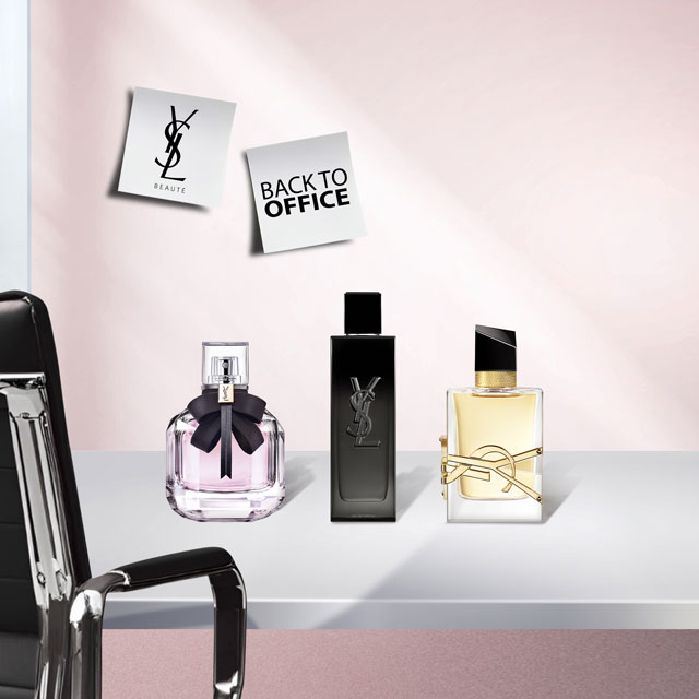 Tre flaconi di profumo YSL (Mon Paris con fiocco nero, YSL Y nero e Libre dorato) posizionati su una scrivania bianca. Una sedia da ufficio nera è visibile a sinistra, e due post-it "YSL BEAUTÉ" e "BACK TO OFFICE" sono appuntati su una parete rosa chiaro.