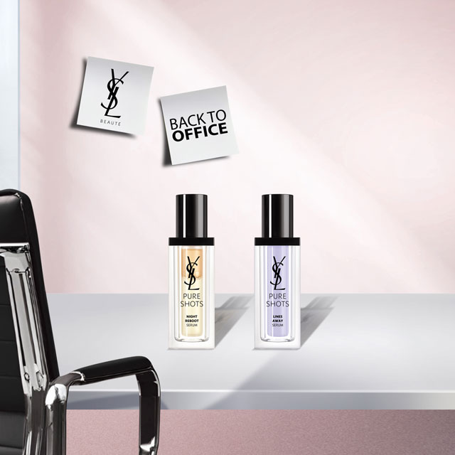 Due flaconi di sieri YSL Pure Shots (un flacone arancione per "Night Reboot" e un flacone viola per "Light Up") posizionati su una scrivania bianca. Una sedia da ufficio nera è visibile a sinistra, e due post-it "YSL BEAUTÉ" e "BACK TO OFFICE" sono appuntati su una parete rosa chiaro.