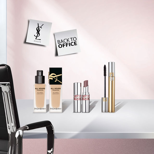 Due flaconi di fondotinta YSL All Hours Foundation Luminous Matte, due tubetti di rossetto YSL e due tubetti di mascara YSL (uno dorato e uno argentato) posizionati su una scrivania bianca. Una sedia da ufficio nera è visibile a sinistra, e due post-it "YSL BEAUTÉ" e "BACK TO OFFICE" sono appuntati su una parete rosa chiaro.