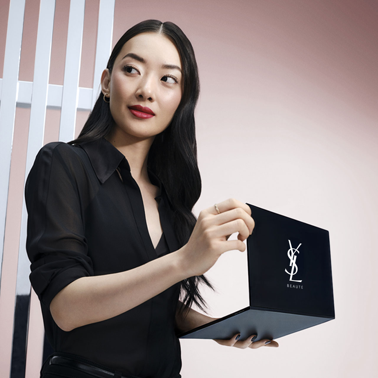 Una donna con lunghi capelli scuri, che indossa una camicia nera, tiene in mano un laptop nero aperto che mostra il logo "YSL BEAUTÉ" in bianco sullo schermo. Guarda verso destra, e lo sfondo presenta un muro rosa chiaro con barre verticali bianche.