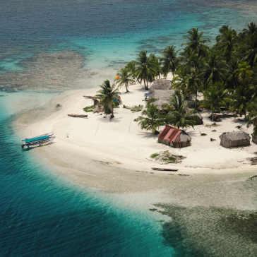 A tropical island with palm trees, huts and turquoise water, as seen from the air.