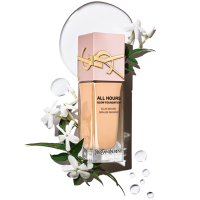Flacone di fondotinta YSL All Hours in una tonalità beige chiaro, posizionato con grazia tra delicati fiori bianchi e goccioline d'acqua scintillanti su una superficie riflettente.
