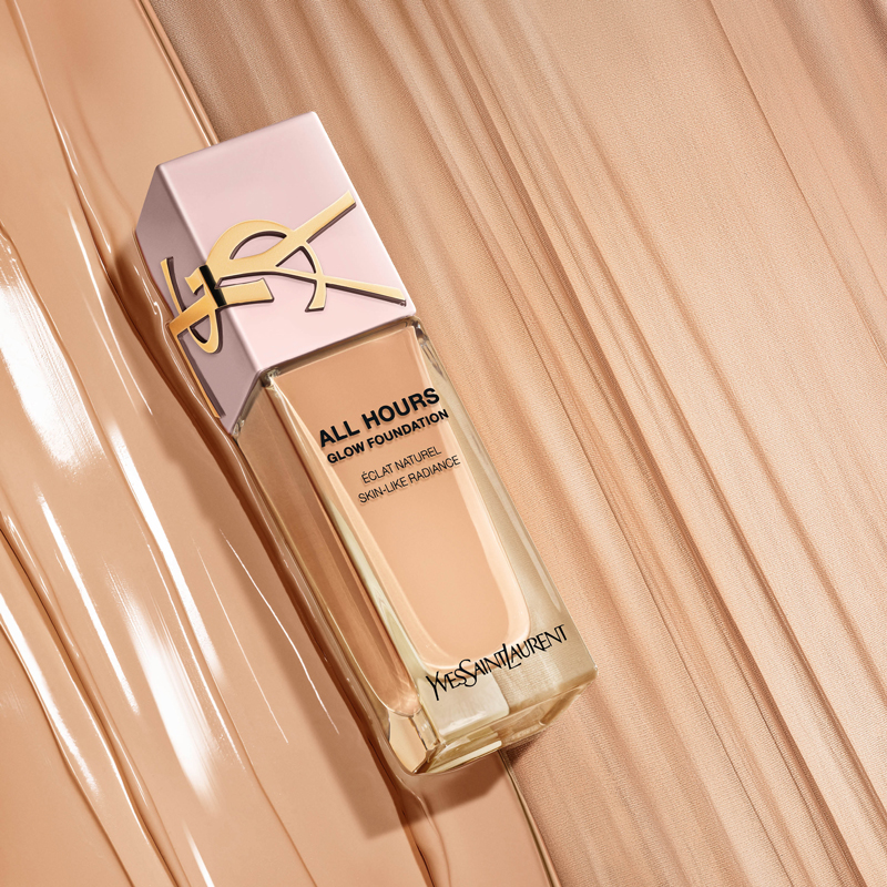 Flacone di fondotinta YSL All Hours in una tonalità beige chiaro, circondato da tracce di fondotinta liquido su una superficie dai toni caldi.