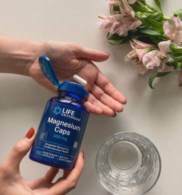 alt="Mani che tengono un flacone Life Extension Magnesium Caps con capsule e un bicchiere vuoto su un tavolo con fiori»