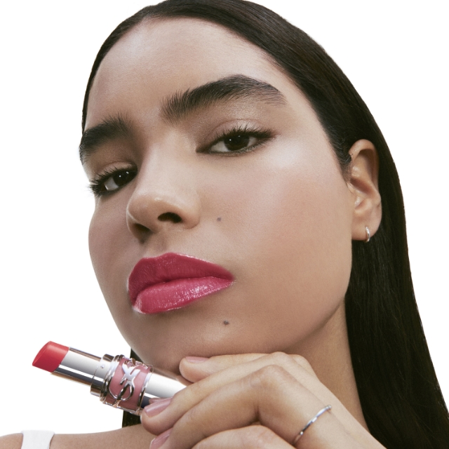 Una donna con capelli scuri e occhi penetranti guarda direttamente la fotocamera, tenendo un tubetto di rossetto YSL aperto color argento e oro rosa vicino al mento. La punta del rossetto è una vibrante tonalità rosa corallo lucida. Ha un anello al dito