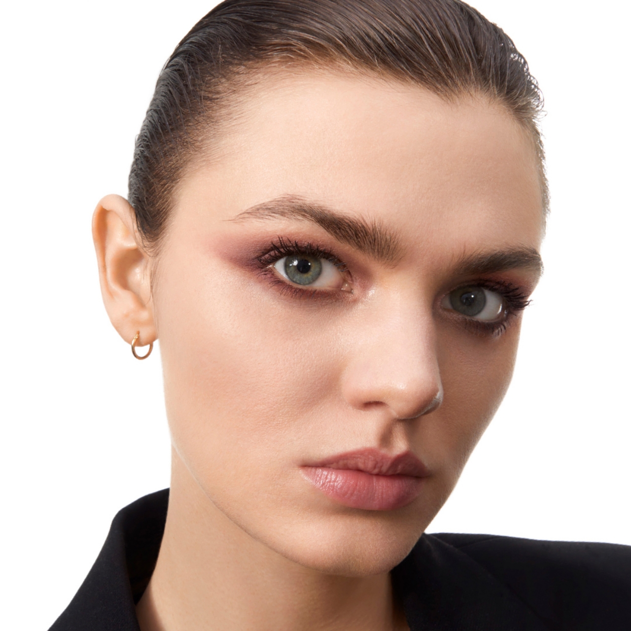 Primo piano del viso di una donna che guarda direttamente la fotocamera, con un audace ombretto rosa e un eyeliner sottile, sopracciglia ben definite e un colore di labbra naturale. Ha gli occhi chiari e un piccolo orecchino a cerchio dorato nell'orecchio sinistro