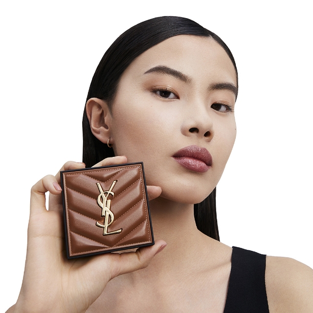 Una donna dai capelli scuri tiene in mano un poudrier quadrato YSL con una finitura trapuntata simile alla pelle marrone e un prominente logo YSL dorato. Ha un trucco occhi delicato e un rossetto marrone rossastro, guardando direttamente la fotocamera. Indossa un piccolo orecchino a cerchio dorato
