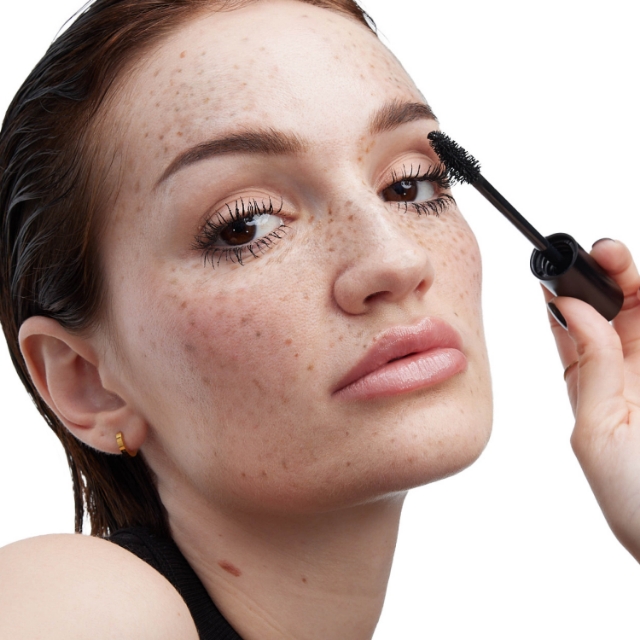 Una donna con le lentiggini è ripresa in primo piano, che guarda leggermente verso l'alto mentre applica il mascara sull'occhio destro con una bacchetta nera. Le sue ciglia appaiono voluminose e ha labbra piene e lucide. Indossa un piccolo orecchino a cerchio dorato