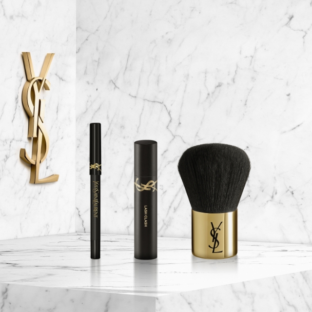 Un grande logo YSL dorato fissato su una parete di marmo bianco, con su una mensola dello stesso marmo, una penna eyeliner nera, un tubetto di mascara nero "LASH CLASH" e un pennello da trucco a setole nere con base dorata e logo YSL