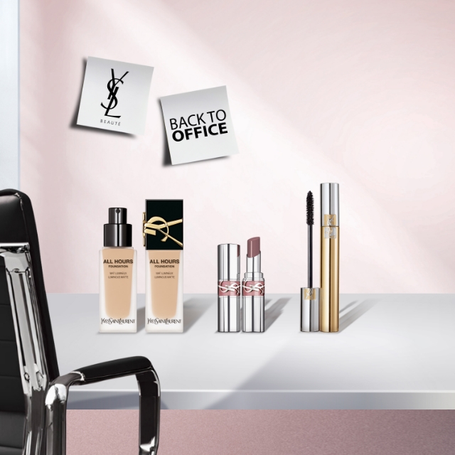 Due flaconi di fondotinta YSL All Hours Foundation Luminous Matte, due tubetti di rossetto YSL e due tubetti di mascara YSL (uno dorato e uno argentato) posizionati su una scrivania bianca. Una sedia da ufficio nera è visibile a sinistra, e due post-it "YSL BEAUTÉ" e "BACK TO OFFICE" sono appuntati su una parete rosa chiaro