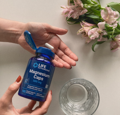 alt="Mani che tengono un flacone di Life Extension Magnesium Caps da 500 mg accanto a un bicchiere d'acqua e fiori rosa»