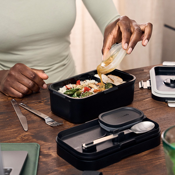 Lunchbox con cibo all’interno su un tavolo, con posate intorno, una persona versa della salsa sul pasto