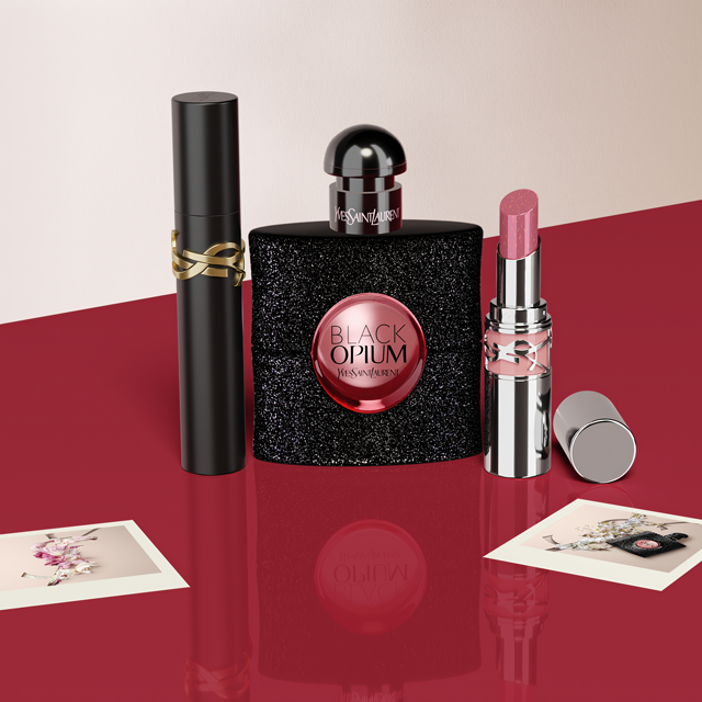 Una bottiglia di profumo nera e scintillante, forse "Black Opium" di YSL, circondata da un rossetto rosso, un cofanetto nero e piccole carte bianche, il tutto disposto su una superficie rossa brillante.