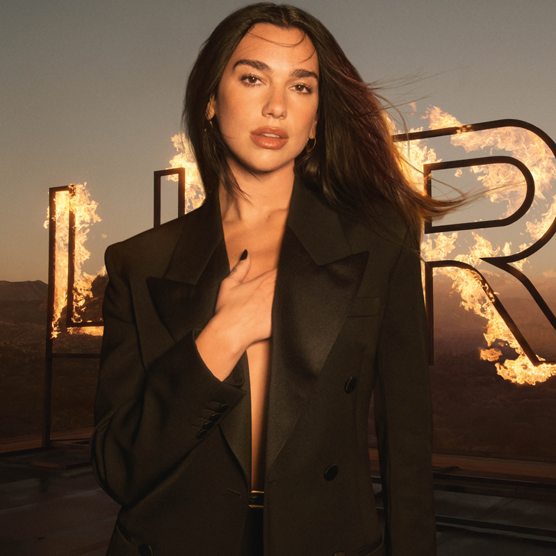 A medium shot of Dua Lipa, an ambassador for Yves Saint Laurent's Libre fragrance, wearing an open black blazer revealing her chest, with her left hand resting over her heart. She stands in front of large, burning letters that spell "LIBRE", set against a sunset or dusk sky.