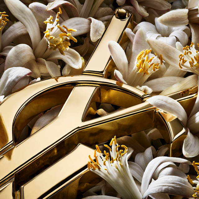 Primo piano del logo YSL dorato e brillante, parzialmente coperto da piccoli e delicati fiori bianchi con stami giallo vivo.