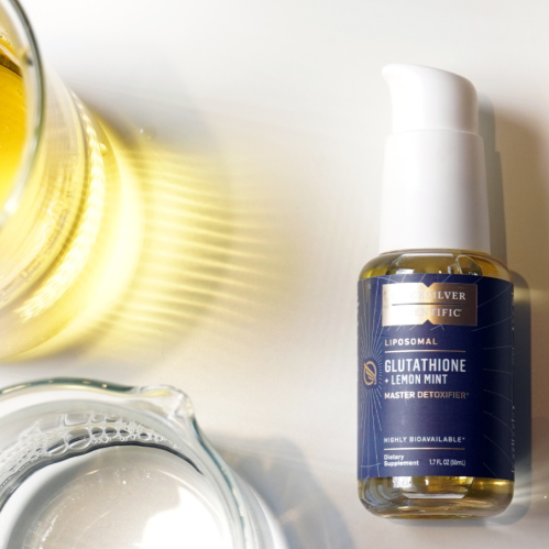 alt="Una bottiglia di glutatione liposomiale adagiata su una superficie bianca accanto a una caraffa d'acqua e un bicchiere d'acqua con gocce di glutatione liposomiale mescolate.»