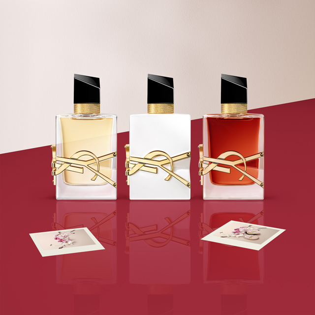 Tre miniature di profumi Yves Saint Laurent (YSL) in bottiglie dorate, arancioni e trasparenti, possibilmente Black Opium, Libre e Mon Paris, esposte su una superficie rossa con piccole carte logo YSL.