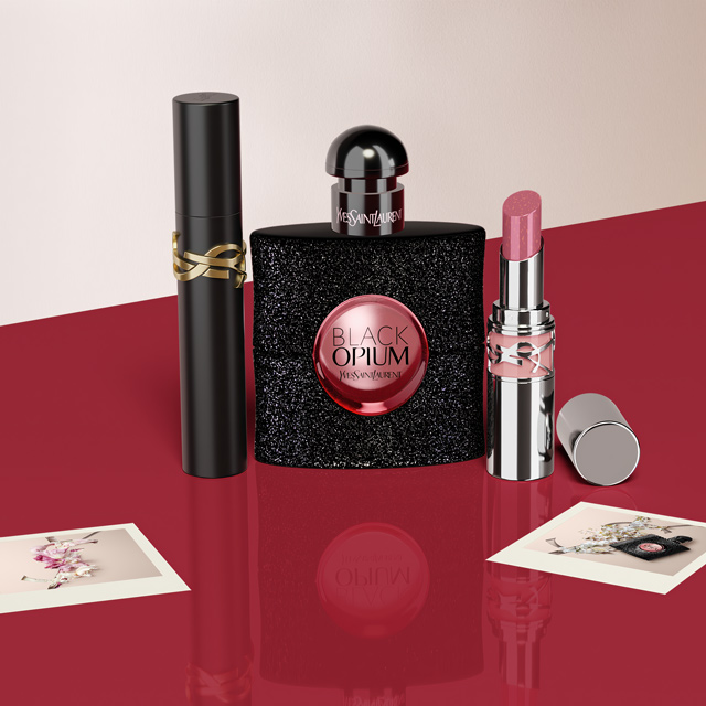 Prodotti di bellezza Yves Saint Laurent (YSL): profumo Black Opium, Mascara Volume Effet Faux Cils e un rossetto Rouge Pur Couture, disposti su una superficie rossa con piccole carte logo YSL.