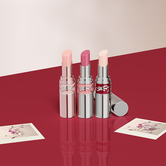 Una selezione di rossetti Yves Saint Laurent (YSL) Rouge Volupté Shine in varie tonalità di rosa e tubi argentati, disposti su una superficie rossa con piccole carte logo YSL.
