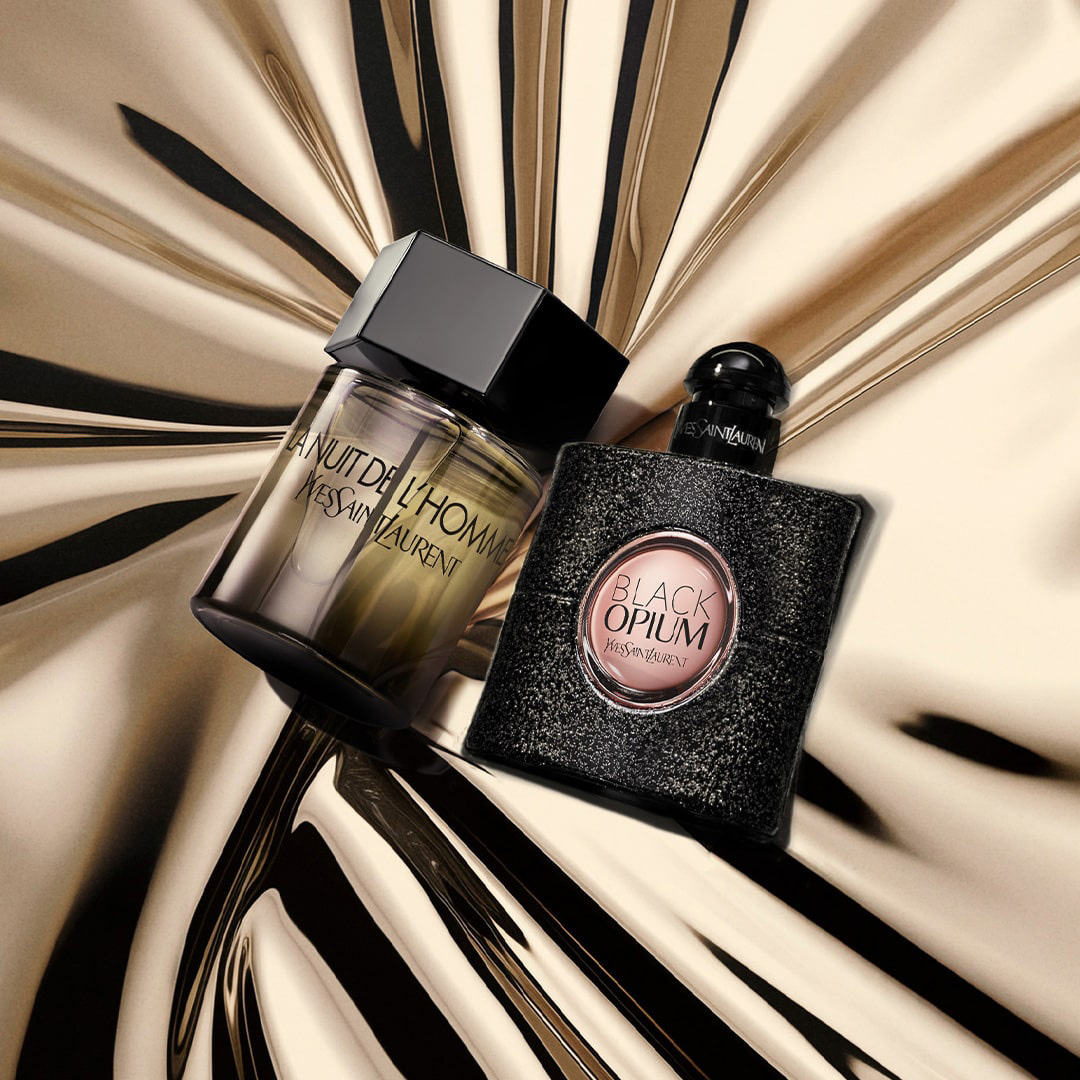 Due iconiche bottiglie di profumo YSL, "La Nuit de l'Homme" e "Black Opium", sono posizionate una accanto all'altra su una superficie dorata e ondulata, creando un elegante riflesso.