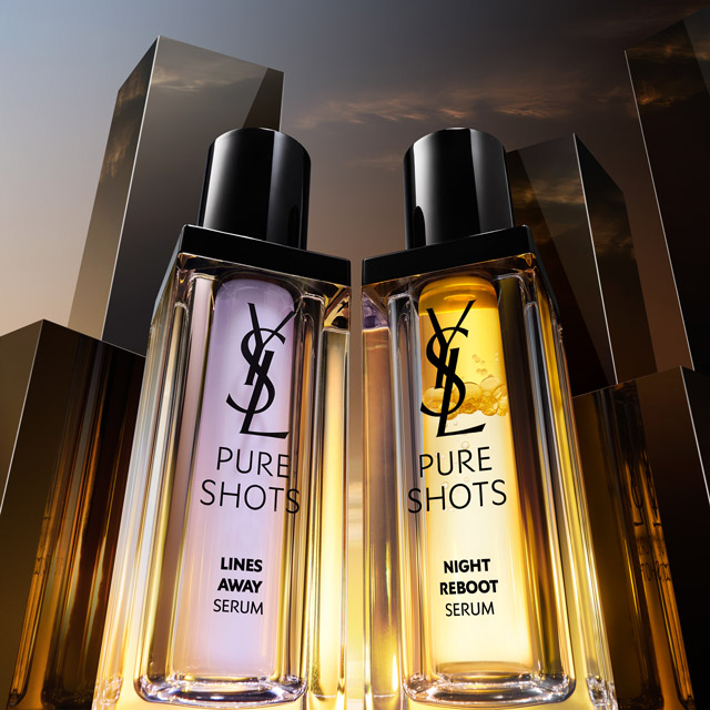 Due sieri YSL Pure Shots, "Lines Away Serum" (viola) e "Night Reboot Serum" (dorato), sono in primo piano su uno sfondo urbano stilizzato con grattacieli scuri e un cielo crepuscolare.