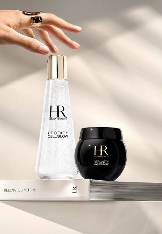 Una mano che si dirige verso il duo Dewy Essence e Age Recovery Night, entrambi appoggiati sullo stesso libro, in una luce morbida.