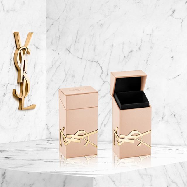Due scatole di profumo rettangolari beige, ornate con il logo YSL dorato, sono posizionate su una superficie di marmo bianco. Una scatola è chiusa mentre l'altra è aperta, rivelando un interno nero. Un grande logo YSL dorato è apposto sulla parete di marmo bianco sullo sfondo