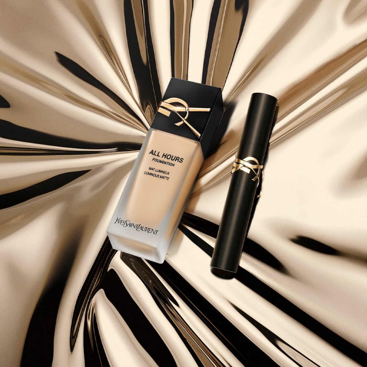 Un flacone di fondotinta "All Hours Foundation" di Yves Saint Laurent e un prodotto cilindrico (probabilmente uno stick) posati su una superficie dorata lucida e ondulata. Il fondotinta è di colore beige chiaro e il suo tappo nero è ornato dal logo YSL dorato