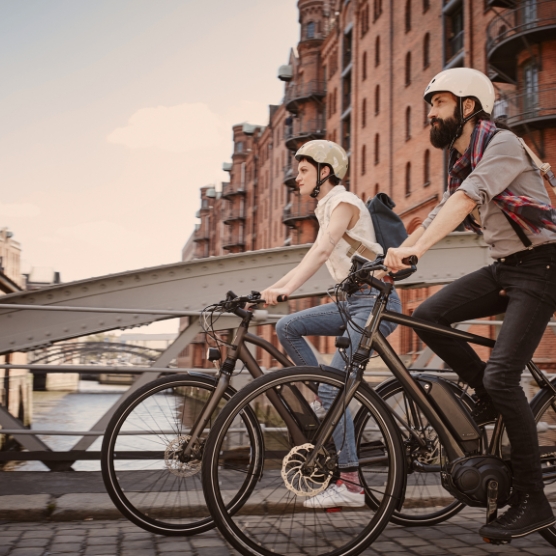 Two people wearing helmets ride bicycles side by side across a bridge, with red brick buildings and a canal in the background.
