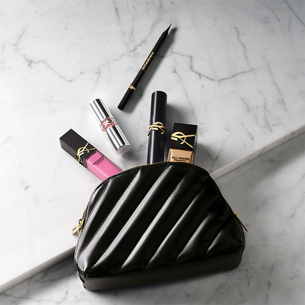Una pochette trapuntata nera è appoggiata su una superficie di marmo, circondata da prodotti di bellezza Yves Saint Laurent tra cui rossetto, fondotinta e eyeliner.