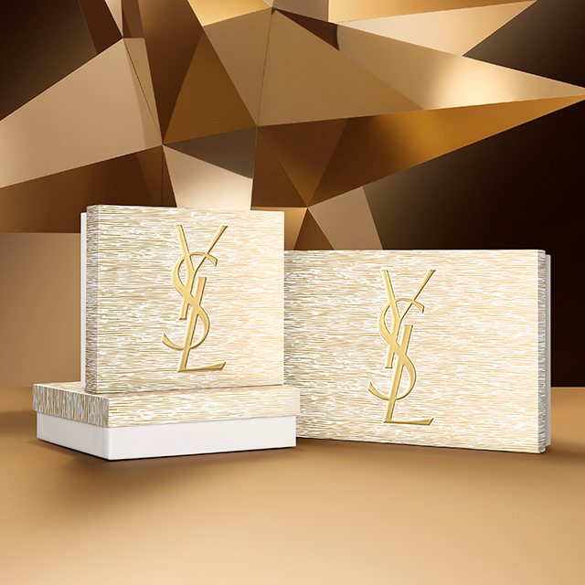 Due eleganti scatole regalo YSL, una più piccola e una più grande, sono ben visibili. Entrambe le scatole sono bianche con un motivo dorato testurizzato e presentano il distintivo logo YSL dorato, su uno sfondo dorato caldo.