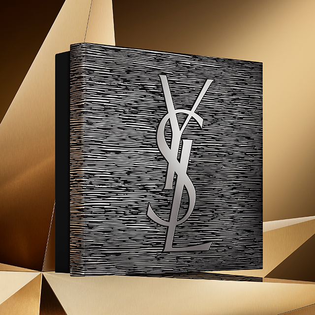 Un'elegante e grande scatola regalo YSL nera, con un motivo testurizzato argentato e un prominente logo YSL argentato, è presentata su uno sfondo dorato con faccette geometriche.