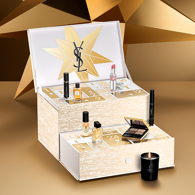 Un calendario dell'Avvento YSL aperto rivela numerosi scomparti, ognuno contenente un prodotto di bellezza in miniatura. Il calendario è bianco con motivi dorati e una grande stella dorata sul coperchio, circondato da altri prodotti YSL come una candela, un profumo e del trucco.
