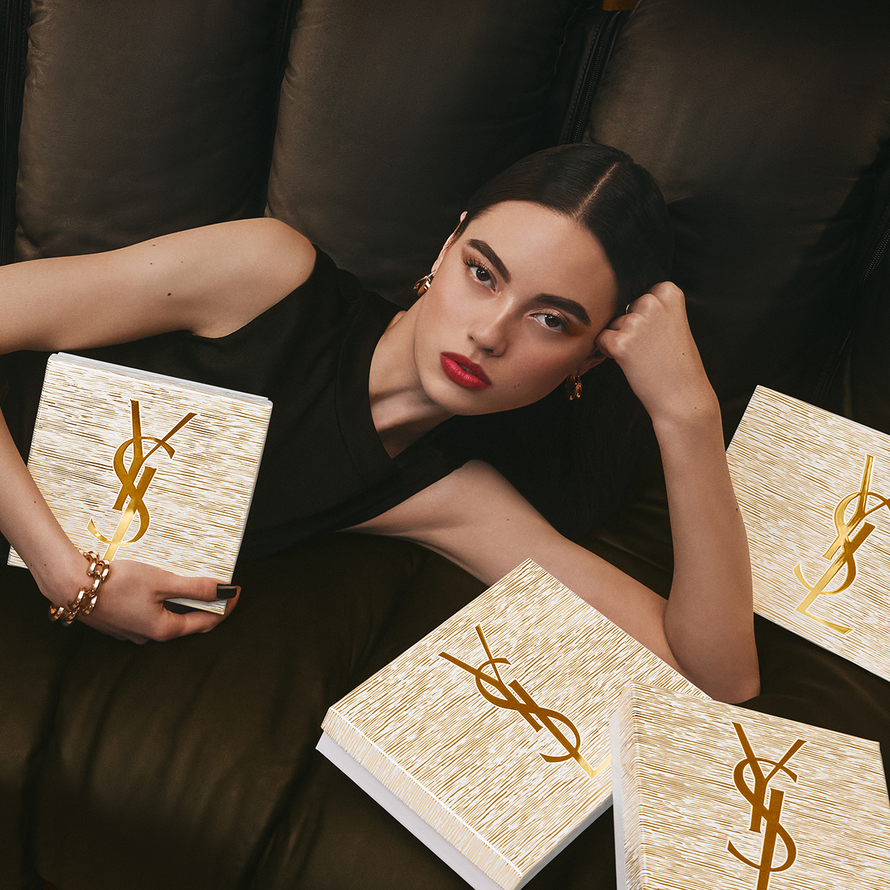 Una giovane donna con capelli scuri e un trucco suggestivo, incluso il rossetto rosso, è sdraiata su un divano in pelle marrone scuro. Tiene una scatola regalo YSL bianca con logo dorato, e diverse altre scatole abbinate sono posizionate intorno a lei.