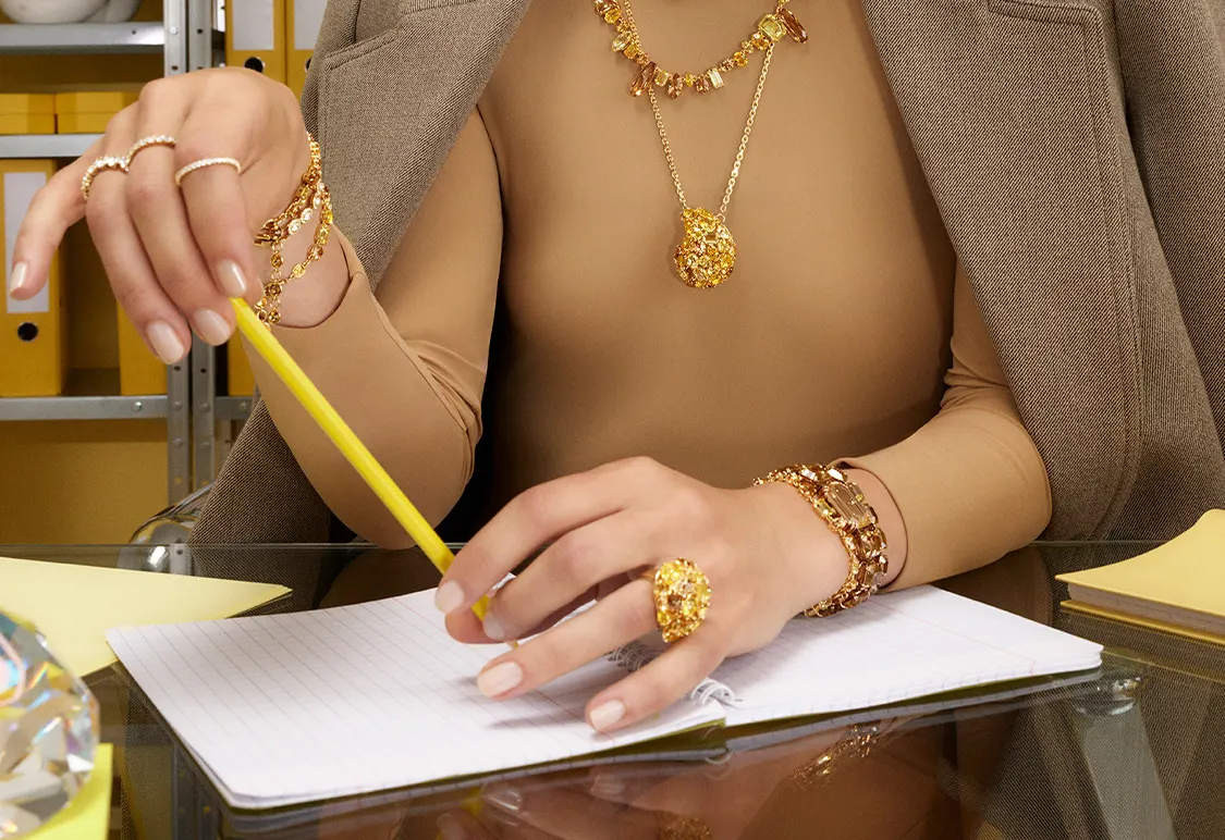 La modella indossa gioielli Swarovski in tonalità oro seduta a una scrivania con un blocco di carta e con in mano una penna gialla