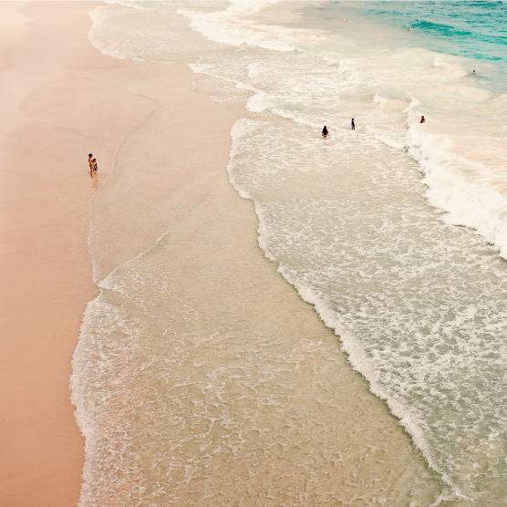 Top view of a wide sandy beach with gentle waves rolling ashore. On the left, a person is walking along the waterline, while on the right, several people are in the shallow sea.