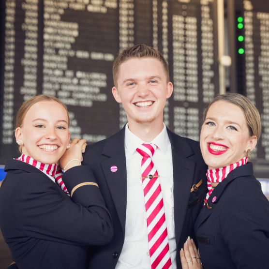 Three flight attendants in uniform stand close together and smile at the camera. Two women wear a pink-and-white striped neck scarf, and the man wears a matching tie. In the background, a large departure board with flight information is visible.If image islinked, link description goes here - otherwise delete this text