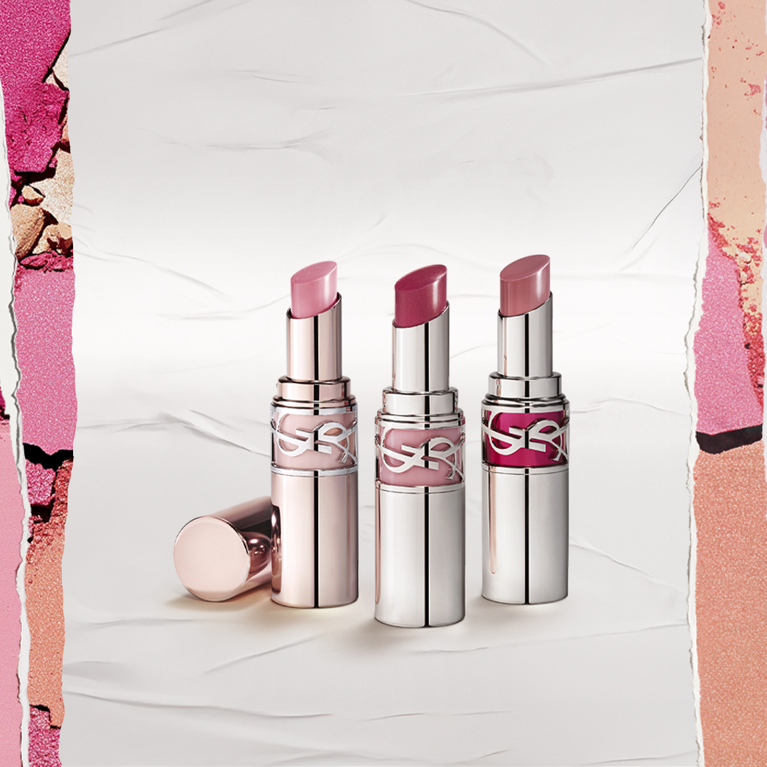 Tre rossetti YSL Rouge Volupté Shine, ciascuno nell'astuccio color oro rosa, sono elegantemente in piedi su una superficie bianca dai bordi strappati, mostrando una gamma di vivaci tonalità rosa e fucsia su uno sfondo di ombretto in polvere.