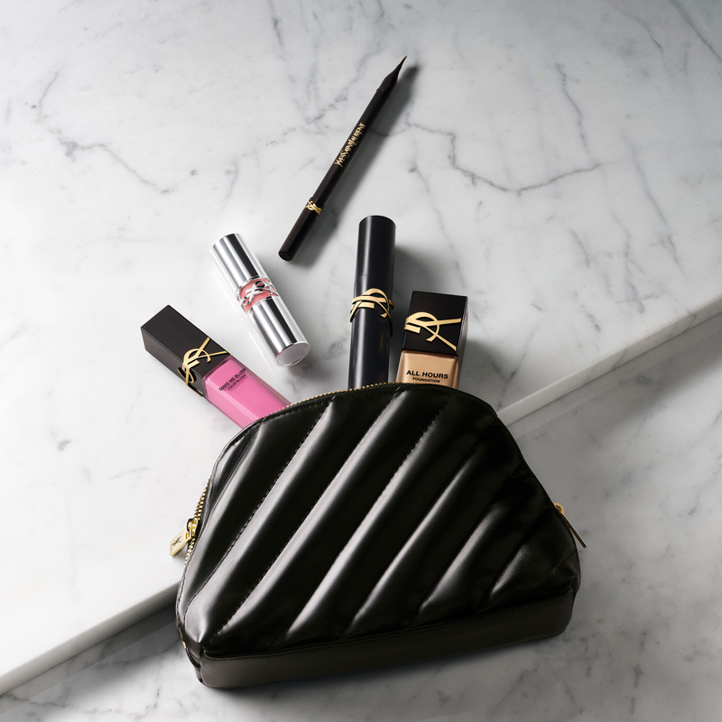 Una lussuosa pochette trucco YSL nera trapuntata aperta su un'elegante superficie in marmo, che rivela un assortimento di prodotti essenziali YSL Beauty, tra cui fondotinta, rossetto, tinta labbra liquida e un eyeliner.