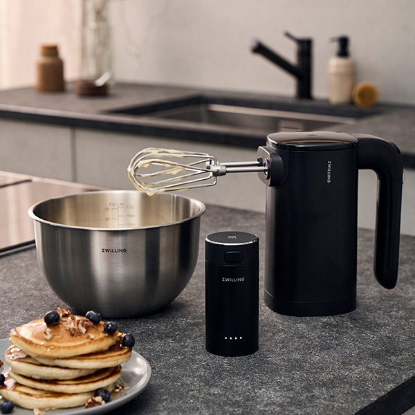 Frullatore a mano sul piano cucina, con pancake in primo piano e una ciotola sullo sfondo