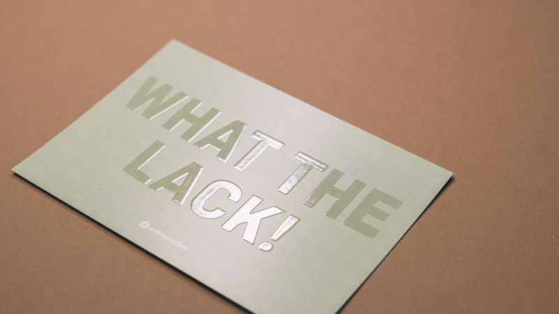 Biglietto beige con scritta in lettere maiuscole argentate “WHAT LACK!” su sfondo marrone chiaro.
