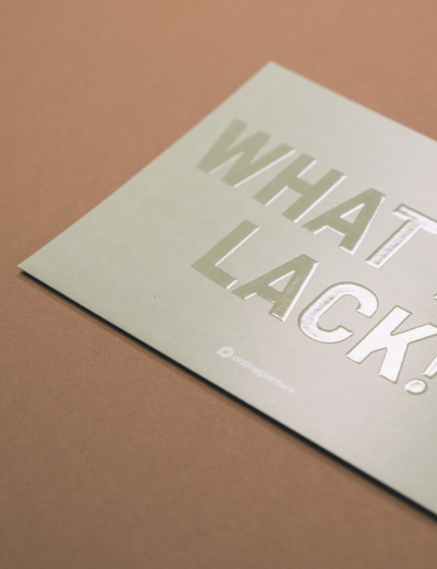 Biglietto beige con scritta in lettere maiuscole argentate “WHAT LACK!” su sfondo marrone chiaro.