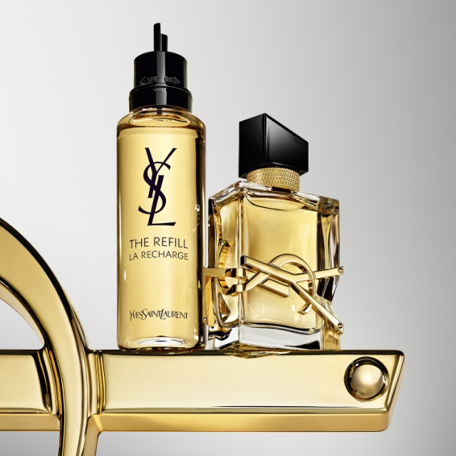 La distintiva bottiglia del profumo YSL Libre è affiancata dalla sua bottiglia di ricarica corrispondente, entrambe con tonalità dorate e l'iconico logo YSL, posizionate su una base decorativa dorata